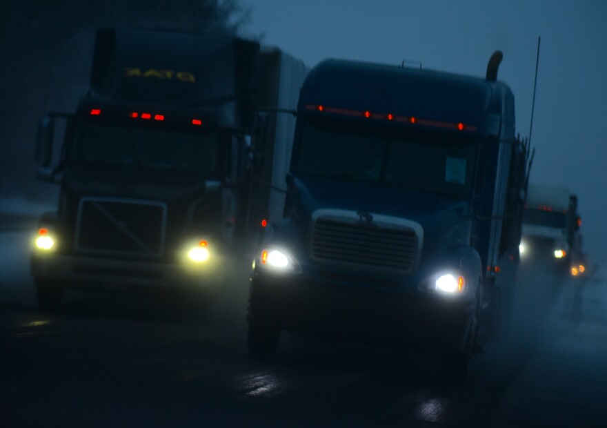 A group of semi-trucks drive toward the camera down a highway in partial darkness. Their headlights are shining as they kick up what appears to be a mist in the road behind them.