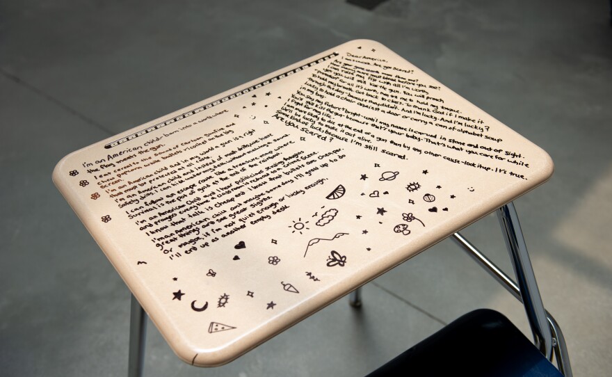 Artwork by Sereen Amezzane of a student desk with text and drawings on it. 