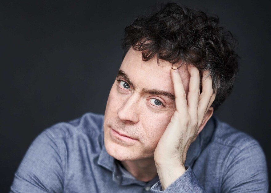 Paul Lewis sits in front of a charcoal background in a blue button-up. He rests his head on his palm and looks at the camera, smoldering. He has dark blue eyes and curly, black hair.