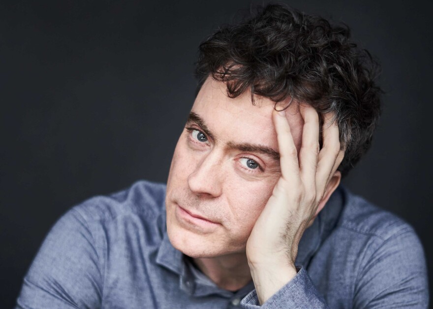 Paul Lewis sits in front of a charcoal background in a blue button-up. He rests his head on his palm and looks at the camera, smoldering. He has dark blue eyes and curly, black hair. 