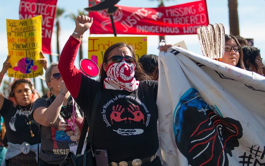 A woman wearing a black t-shirt, sunglasses and red and white bandana covering her nose and mouth holds a white banner in one hand and raises a fist with the other while other people hold signs up behind her.