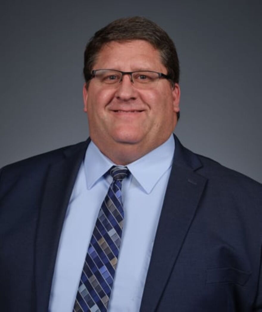 A friendly man is smiling. He wears glasses and has on a blue suit, lighter blue shirt and matching geo-print tie.