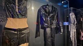 Two stage outfits hang side by side in a glass museum case: a black leather vest and skirt with chain lacing, and a black leather jacket with metal-studded pants. On the right, a silver and denim stage outfit hangs in another glass museum case.
