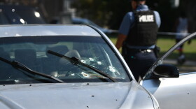 A police officer with his back turned to the camera walks away from a silver car that has seven bullet holes in the windshield and several other bullet holes in the body of the car.