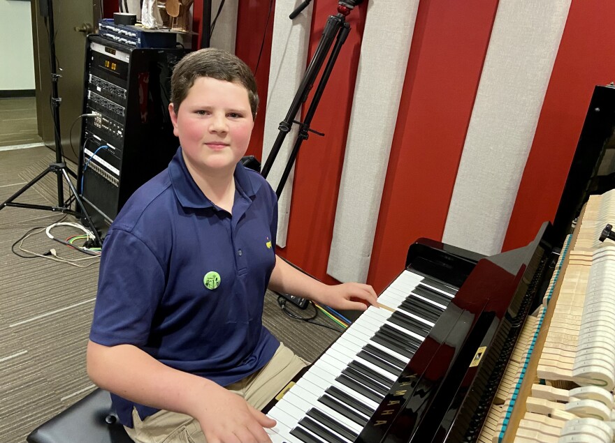 A boy with short brown hair wearing a blue polo shirt sits at a piano in a music studio.