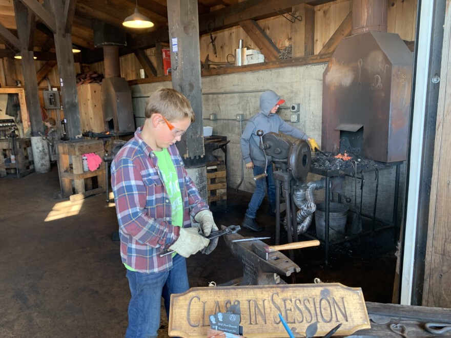 Boy attends to coal burning forge in background while another camper works on his project at an anvil.