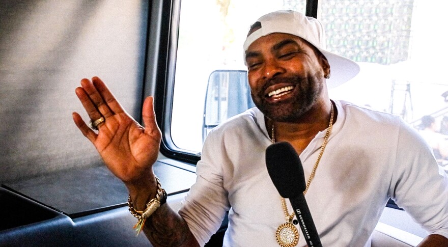 Ginuwine laughs with his hand up in the passenger seat of a trailer. He is wearing a gold chain, a ring and bracelets with an all-white fit.