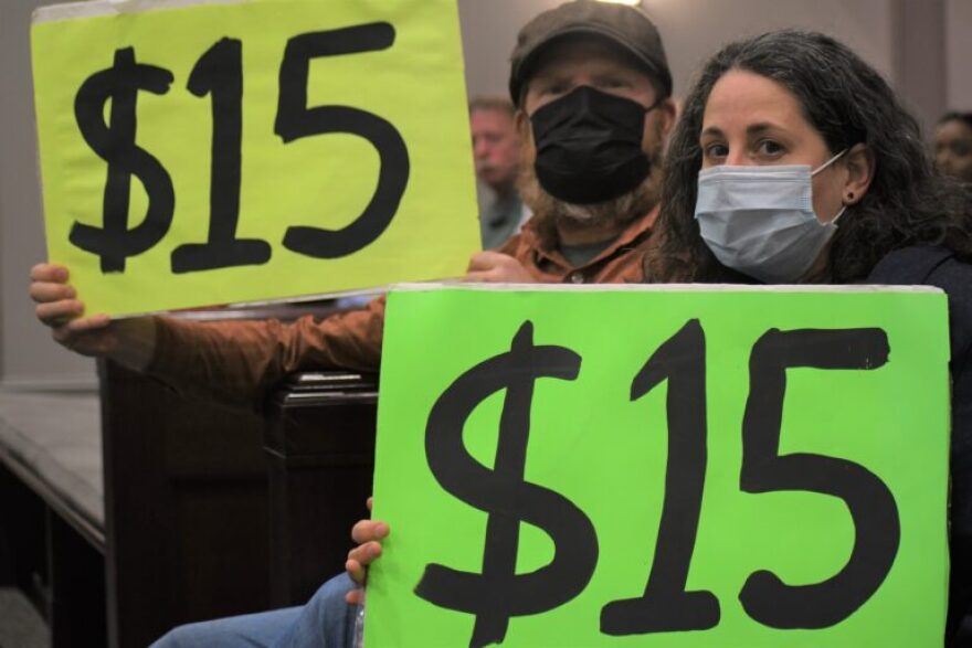 Two people seated in City Hall chambers hold posters with "$15" written on them