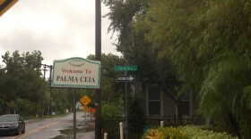 A vintage sign reads "Welcome to PALMA CEIA" on a street corner. Lush greenery, one house and one car are visible in the background.