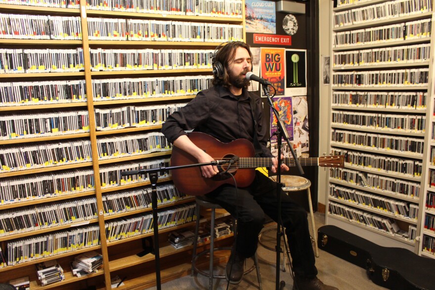 A seated man playing guitar and singing into a microphone in a radio studio with walls lined with shelves of CDs.