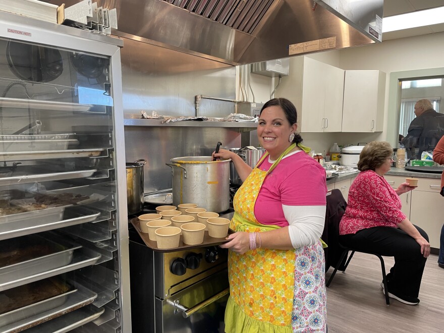 A woman in a pink shirt and yellow apron holds a ladle in a large pot. She's standing in an industrial kitchen and balancing a few small bowls on a tray on the stove. 