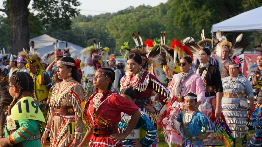 More than 20 Native American are shown dancing in traditional clothing during a powwow. They are wearing their hair in braids and some have feather headdresses on.