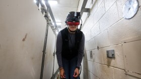 A person wearing a specialized virtual reality headset bends forward while standing in a narrow, indoor corridor with white walls.