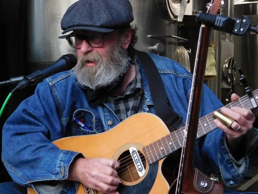 A bearded man playing an acoustic guitar and singing into a microphone.