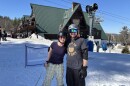 Two skiers in t-shirts pose for a photo in front of a ski lodge. 