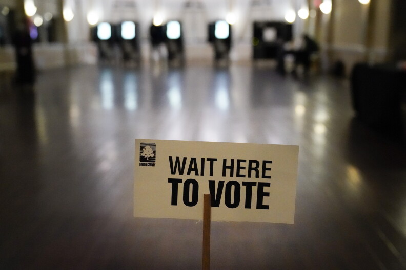 A white sign that reads "Wait here to vote" in all black, capital letters is in the foreground. Voting booths are out of focus along the back pf the image.