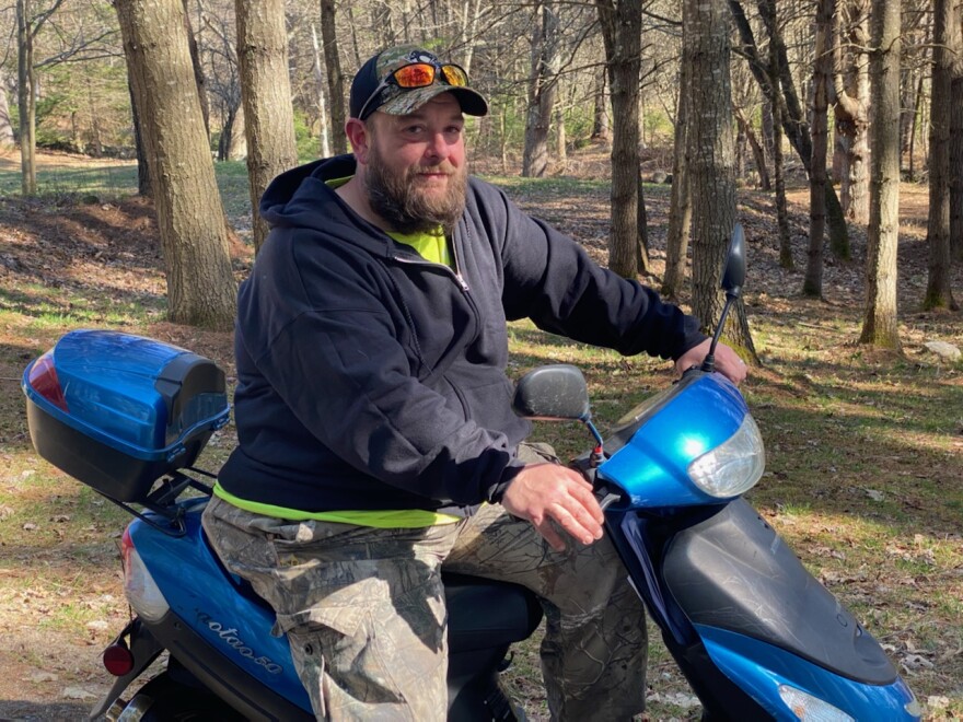 A photo of a man on a blue moped, posing with many trees in the background. 