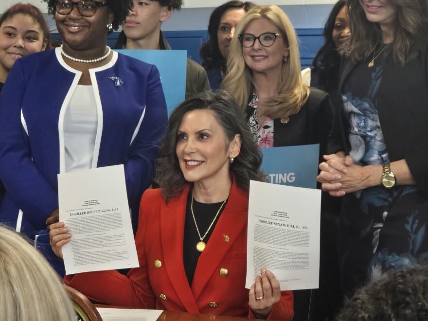 Governor Gretchen Whitmer sits at a desk and holds up two printed documents. She smiles. She wears a bright red blazer with gold buttons.