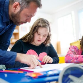 Mane leaning to point at workbook, with two students sitting at table looking at the booklets
