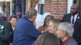 An outdoor campaign gathering where several people interact in front of a brick building. In the center, a man wearing a blue fleece jacket stands with his back partially to the camera, greeting and interacting with a smiling woman with blonde hair and glasses. Other smiling onlookers surround them. Some individuals in the crowd wear campaign buttons that read "Mike Rogers." In the background, a shop window displays colorful clothing, and white campaign balloons are partially visible.