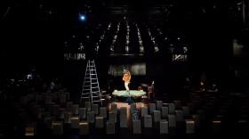 A woman performs on a small circular stage holding an acoustic guitar while surrounded by speakers and an A-frame ladder in an otherwise-empty theater.