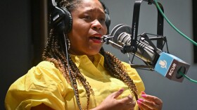 A woman wearing a bright yellow dress gestures with both hands while talking inside a studio at a microphone.