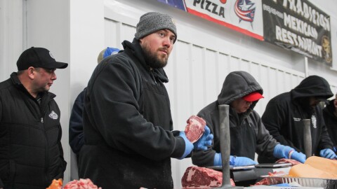 A man holds a raw cut of steak while a larger slab of meat sits in front of him. On the table is a large meat tenderizer and a few scales. Others work around him. 
