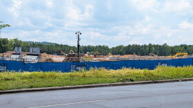 A construction site is visible above a blue privacy fence. Concrete pilings, yellow construction machines and a security camera are visible.