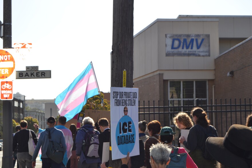 You can see the backs of protestors as the group crosses Baker street. Someone with short gray hair holds up a sign that says "Stop our private date from being stolen!" There's an image of Trump in a racoon mask holding up a big ball that says "ICE Database". A trans flag waves in the front of the crowd. The DMV offices sign is visible in the top right corner of the picture. 