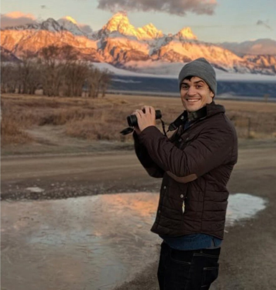 A man with camera smiles in front a blurred mountainous backdrop.