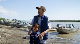 A man and a girl stand on a river shore with boats in the background.