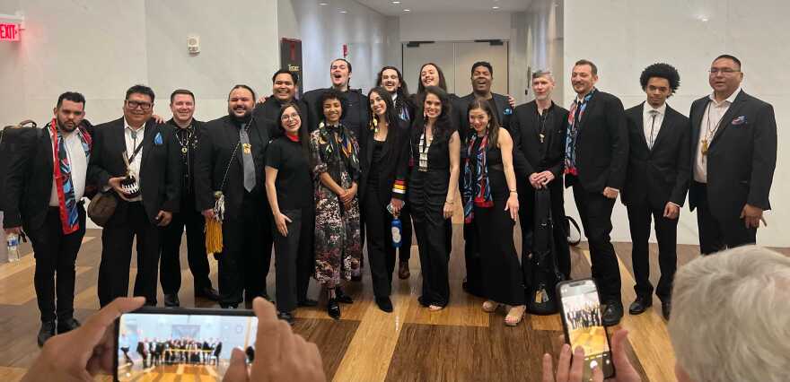 A photo of 18 people posing for a photo and wearing black and various pops of color in scarves, ribbons and pocket squares. In the foreground are hands of people making photos on phones. Everyone is standing on a wood paneled floor, in a room with white walls.