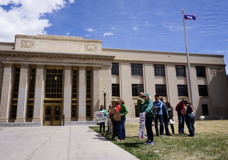 People stand with signs outside a tall cream building with pillars, amid a blue sky.