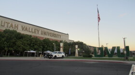 Statues of large green letters read "UVU" in front of a flagpole. There is a building with a sign that reads "Utah Valley University", there is a cop car in front of it. 