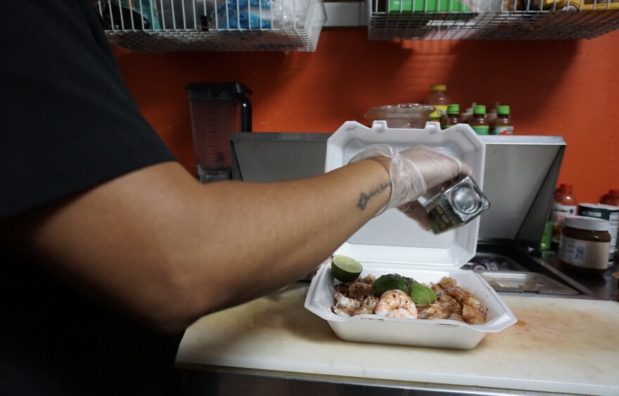 A hand is shown seasoning food in a to-go box filled with shrimp and avocado.