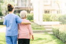 woman in blue scrubs has her arm an older woman with white hair as she escorts her over a grassy green courtyard area, both faces turned away from camera