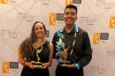 Two dressed-up people hold gold statues and smile at the camera, in front of a backdrop with the words “Heartland Emmy Awards” repeated across the background.