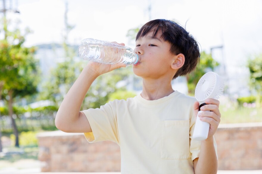 Child drinks water from a plastic bottle while using a portable handheld fan outdoors during a heatwave. 