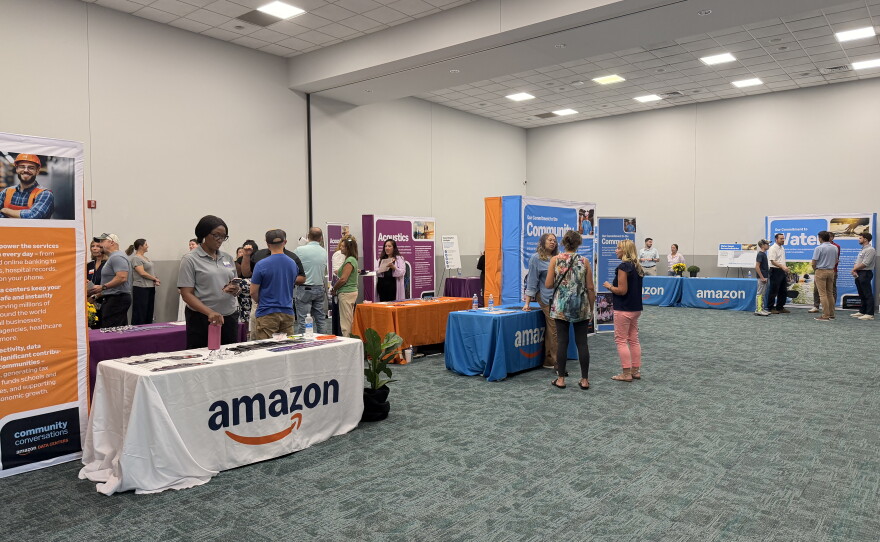 A large room with tables and banners. To the left, there is a woman standing in front of a small table with a white table cloth bearing the Amazon logo. Further down from this table, is another one with a blue table cloth with the Amazon logo. There are three people standing in front of this blue table talking. 