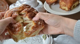 A pair of hands pulls apart a thick cinnamon roll so the viewer can see the inside of a baked cinnamon roll. In the background are other cinnamon rolls of varying shapes, sizes and styles.