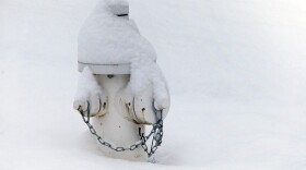 A white fire hydrant is covered in and surrounded by snow. There are chains on the caps.