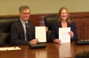 A grey haired man with glasses and a suit holds up a piece of paper. Next to him a blond haired woman holds up a piece of paper. 