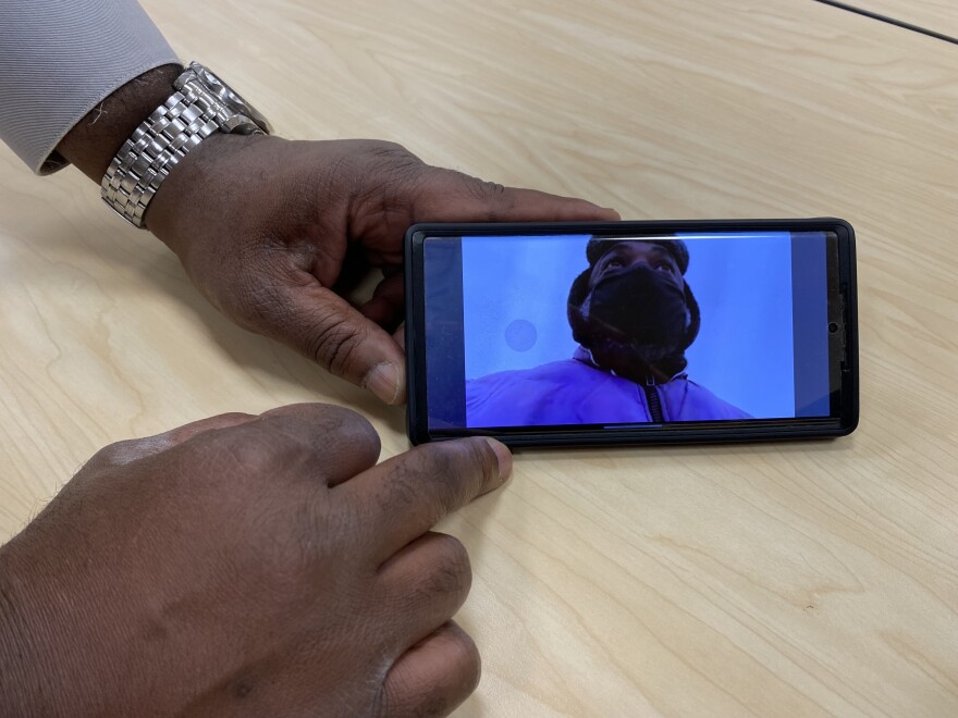 A close-up of a man's hands, pointing at the screen of a phone. The phone screen shows the same man dressed up warmly for winter and wearing a face mask.