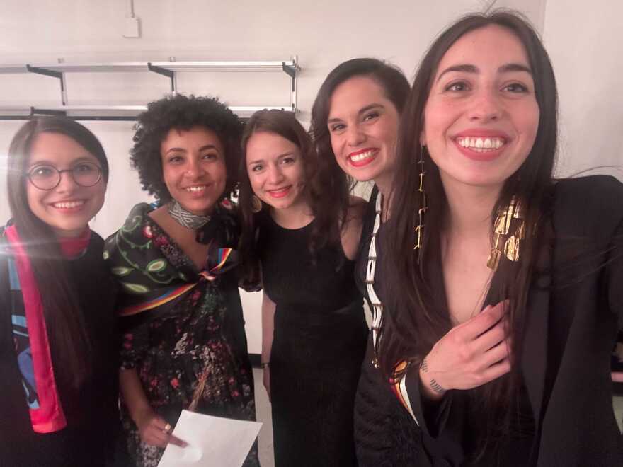 A photo of five femmes crowded together and smiling at the camera. 