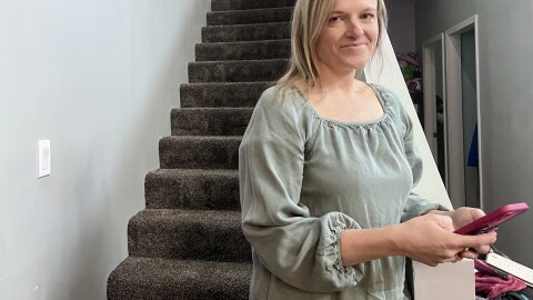 A woman stands at the base of the stairs inside a home as she holds a phone. 