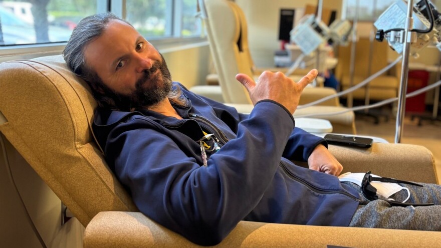 man with dark hair and full beard is lying down on a recliner with IV tubes and gives a hand gesture on thumb and pinky up to acknowledge he is fine 