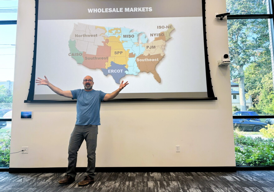 A man stands with arms outstretched in front of a screen showing a map of U.S. wholesale energy markets in a modern, bright room with large windows.
