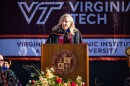 A woman with blonde hair stands at a podium, wearing a black robe with a maroon sash. 