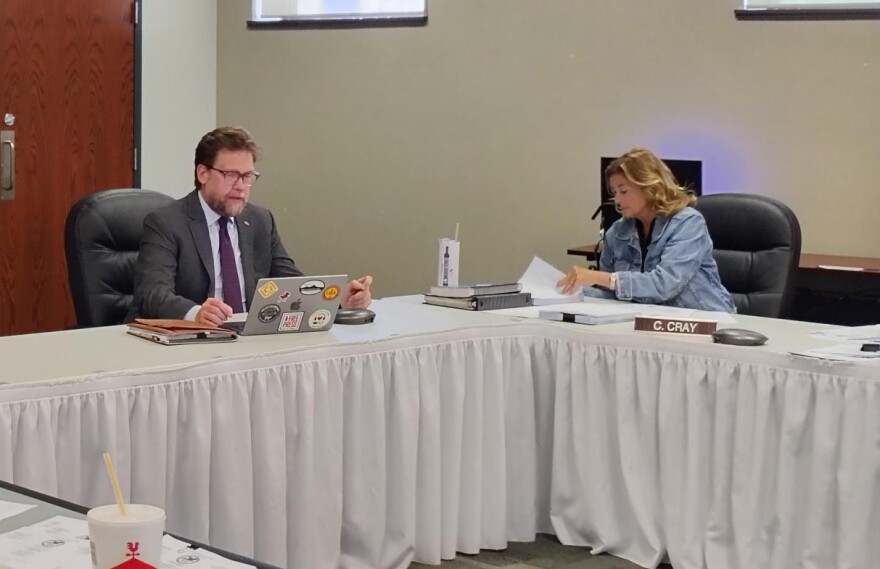 Three people sit at a conference table in a meeting room. One man types on a laptop, while two women review documents. Papers, folders, and a drink cup are on the table. The room has beige walls and windows.
