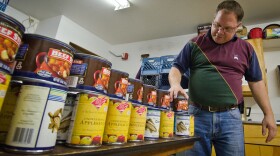 A man in a red and green shirt with blue jeans is standing next to two rows of canned food that's sitting on a table. He is in a small white room with fluorescent lights above him.